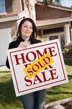 Happy attractive hispanic woman holding sold home for sale sign in front of house