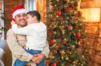 Hispanic armed forces soldier wearing santa hat hugging son in front christmas tree