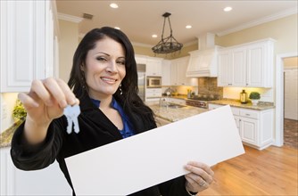 Pretty hispanic woman in kitchen holding house keys and blank white sign