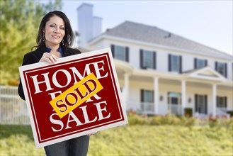 Smiling hispanic woman holding sold home for sale sign in front of beautiful house
