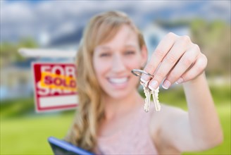 Excited woman holding house keys and sold real estate sign in front of nice new home