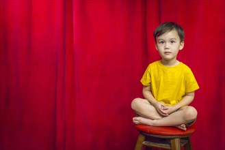 Handsome mixed-race boy sitting on stool in front of red curtain
