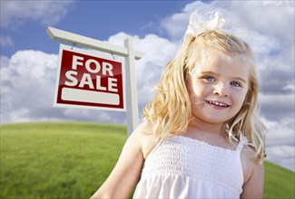 Adorable smiling girl in grass field with for sale real estate sign behind her