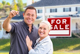 Young adult couple with house keys in front of home and for sale real estate sign