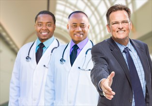 Two mixed-race doctors behind businessman reaching for a hand shake inside hospital