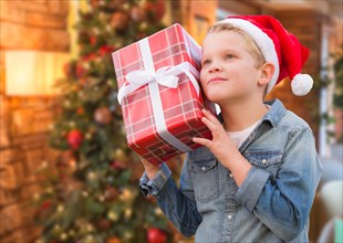 Curious young boy wearing santa hat holding christmas gift in front of decorated tree