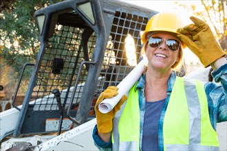 Smiling female worker holding technical blueprints near small bulldozer at construction site