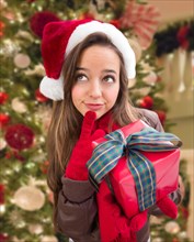 Thinking girl wearing A christmas santa hat with bow wrapped gift in front of decorated tree