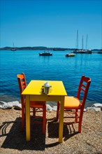 Yellow cafe restaurant table of street cafe with chairs on beach in Adamantas town on Milos island with Aegean sea with boats and yachts in background. Milos island