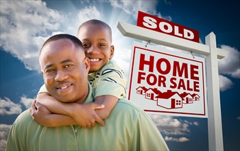Happy african american father with son in front of sold home for sale real estate sign and sky