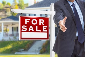 Male agent reaching for hand shake in front of for sale sign and house