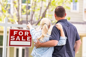Caucasian couple facing and pointing to front of for sale real estate sign and house