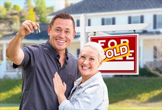 Young adult couple with house keys in front of home and sold for sale real estate sign