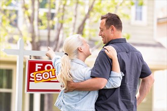 Caucasian couple facing and pointing to front of sold real estate sign and house