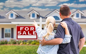 Young adult couple facing and pointing to front of for sale real estate sign and house