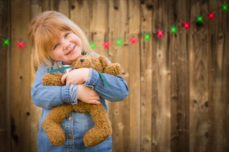 Cute little girl holding her teddy bear in front of wooded background with christmas lights