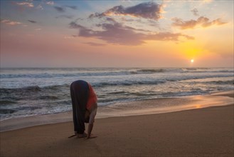 Young sporty fit man doing yoga Sun salutation Surya Namaskar pose Hasta Uttanasana on tropical beach on sunset. With lens flare
