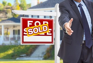 Male agent reaching for hand shake in front of sold for sale sign and house