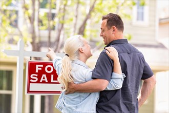 Caucasian couple facing and pointing to front of for sale real estate sign and house