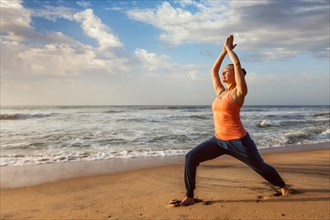 Woman doing Hatha yoga asana Virabhadrasana 1 Warrior Pose outdoors on ocean beach on sunset. Kerala