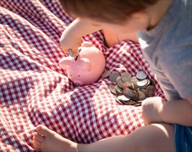 Baby boy sitting on picnic blanket putting coins in piggy bank