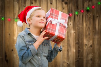 Curious young boy wearing santa hat holding christmas gift on A wood fence background