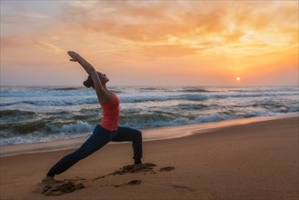 Woman doing Hatha yoga asana Virabhadrasana 1 Warrior Pose outdoors on ocean beach on sunset. Kerala