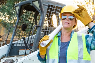 Female worker holding technical blueprints near small bulldozer at construction site