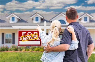 Young adult couple facing and pointing to front of sold real estate sign and house