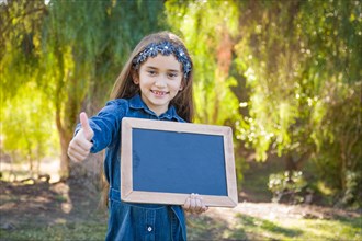Cute young mixed-race girl with thumbs up holding blank blackboard outdoors