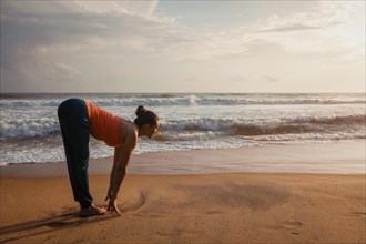 Young sporty fit woman doing yoga Sun salutation Surya Namaskar pose Uttanasana on tropical beach on sunset. With lens flare