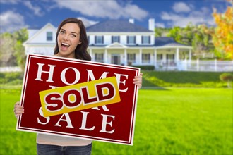 Excited mixed-race female with sold home for sale real estate sign in front of beautiful house