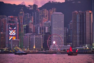 Tourist junk boat ferry with red sails and Hong Kong skyline cityscape downtown skyscrapers over Victoria Harbour in the evening. Hong Kong