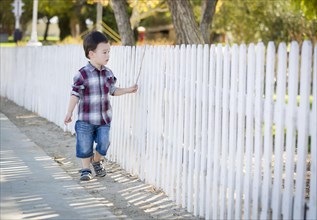 Cute young mixed-race boy walking with stick along white fence
