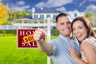 mixed-race excited military couple in front of new home with new house keys and sold real estate sign outside