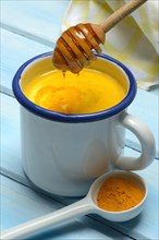 Kurkuma-Milch in Tasse und Honigloeffel mit herablaufendem Bienenhonig und Kurkumapulver