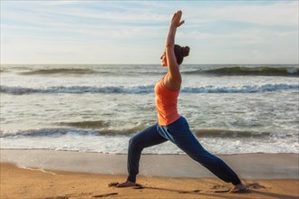 Woman doing Hatha yoga asana Virabhadrasana 1 Warrior Pose outdoors on ocean beach on sunset. Kerala