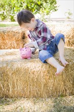 Cute young mixed-race boy sitting on hay bale putting coins into pink piggy bank
