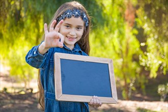 Cute young mixed-race girl with okay sign holding blank blackboard outdoors