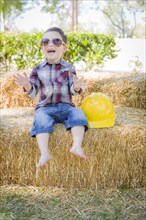 Cute young mixed-race boy laughing with sunglasses and hard hat outside sitting on hay bale