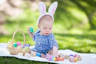 mixed-race chinese and caucasian baby boy outside wearing rabbit ears playing with easter eggs