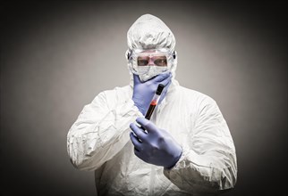 Man wearing HAZMAT protective clothing holding test tube filled with blood against A gray background