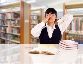 Young female mixed-race student stressed and frustrated in library with blank pad of paper and books