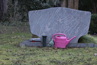 Pinkfarbene Giesskanne und Laubharke hinter einem Grabstein