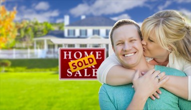 Happy couple hug in front of sold real estate sign and beautiful new house