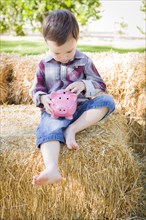 Cute young mixed-race boy sitting on hay bale putting coins into pink piggy bank