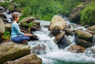 Woman in Hatha yoga asana Padmasana outdoors at tropical waterfall