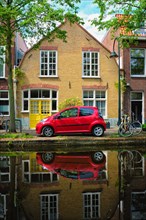 Red car on canal embankment in street of Delft with reflection and bicycle. Delft