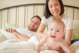 Young mixed-race chinese and caucasian baby boy laying in bed with his father and mother
