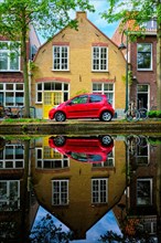 Red car on canal embankment in street of Delft with reflection and bicycle